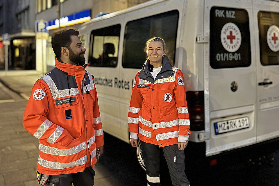 Ein Mann und eine Frau stehen vor einem DRK-Fahrzeug und tragen Rotkreuzjacken.