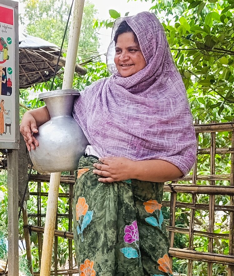 Eine lächelnde Frau in Cox's Bazar, Bangladesch, steht im Freien und hält einen silbernen Wassertopf gegen ihre Hüfte. Sie trägt ein lila Kopftuch und einen grünen Rock mit Blumenmuster, im Hintergrund sind üppiges Grün und Bambusstangen zu sehen.