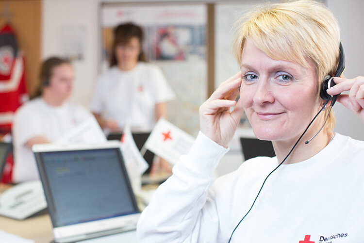 Eine DRK-Mitarbeiterin sitzt vor einem Laptop und trägt ein Headset. Im Hintergrund sind zwei weitere DRK-Mitarbeiterinnen