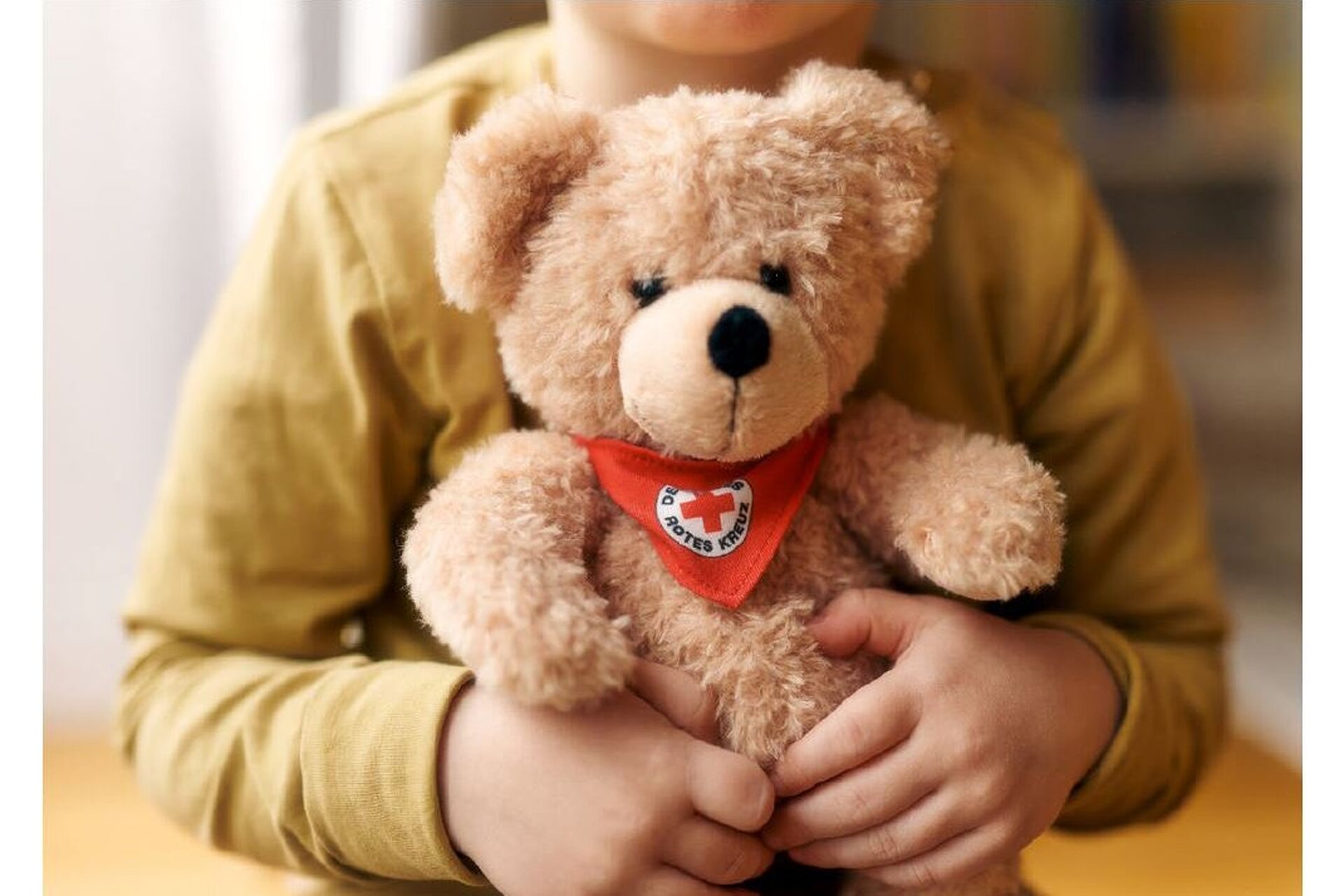 Eine Nahaufnahme von einem etwa vierjährigen Kind. Zu sehen ist lediglich der Oberkörper ohne Kopf, das Kind trägt einen senfgelben Longsleeve und hält einen hellbraunen Teddybär in beiden Händen vor sich. Der Teddybär trägt ein rotes Halstuch mit einem Roten Kreuz auf weißem Kreis und der Aufschrift "Deutsches Rotes Kreuz"