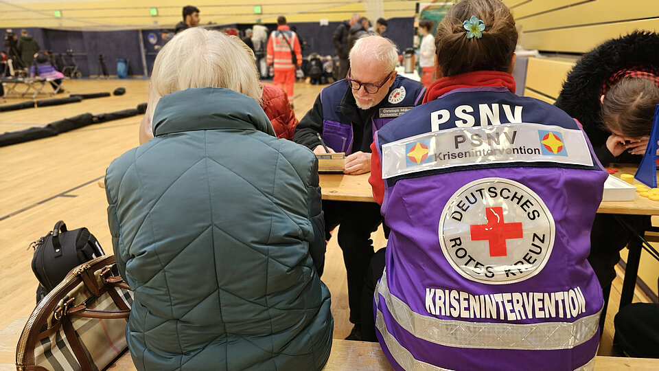Krisenintervention des DRK In einer Turnhalle sitzen Menschen an einem Tisch. PSNV-Einsatzkräfte des DRK in violetten Westen nehmen Daten auf.