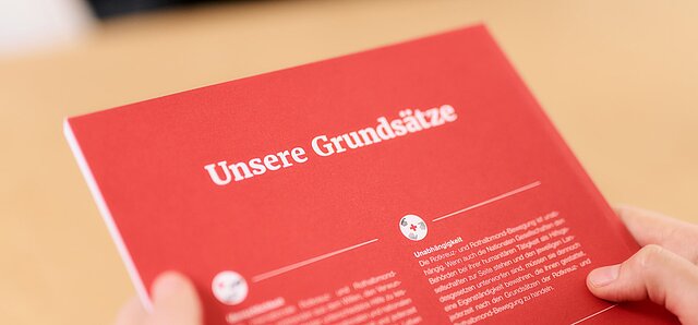 Zu sehen ist ein rotes Heft, auf dem mit weißer Schrift die Grundsätze des deutschen roten Kreuzes und der gesamten Rotkreuz- und Rothalbmonbewegung stehen. Erkennbar ist nur die Überschrift "Unsere Grundsätze" sowie der Text zu Unabhängigkeit: Die Rotkreuz- und Rothalbmondbewegung ist unabhängig. Wenn auch die Nationalen Gesellschaften den Behörden bei ihrer humanitären Tätigkeit als Hilfsgesellschaften zur Seite stehen und den jeweiligen Landesgesetzen unterworfen sind, müssen sie dennoch eine Eigenständigkeit bewahren, die ihnen gestattet, jederzeit nach den Grundsätzen der Rotkreuz- und Rothalbmondbewegung zu handeln.  