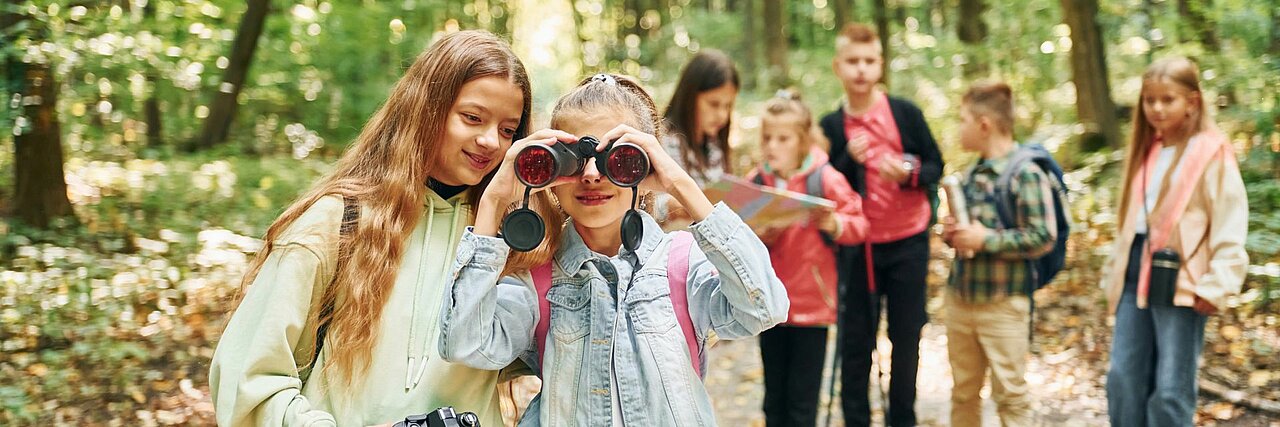 Zwei Mädchen stehen auf einem sonnigen Waldweg. Eines blickt durch ein Fernglas, das andere schaut zu ihr. Im Hintergrund steht eine Gruppe Kinder, sie sind nur verschwommen erkennbar.