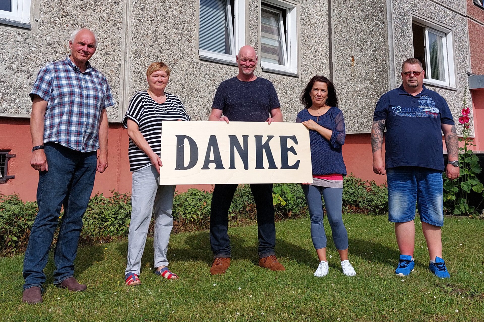 Auf dem Bild stehen fünf Mitarbeiterinnen und Mitarbeiter der Unterkunft für Obdachlose in Stralsund vor ebendieser Unterkunft. Sie stehen auf einer Wiese, das Gebäude im Hintergrund ist hellrot und grau. Die Personen in der Mitte halten ein großes Schild mit der Aufschrift "Danke" in die Kamera.