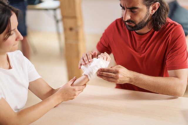 Erste Hilfe Situation: Versorgung einer Schnittwunde an der Hand, die sich eine Frau beim Gemüseputzen in der Küche zugezogen hat. Der Mann legt eine Mullbinde an.