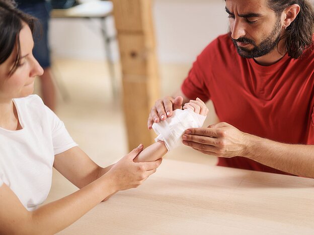 Erste Hilfe Situation: Versorgung einer Schnittwunde an der Hand, die sich eine Frau beim Gemüseputzen in der Küche zugezogen hat. Der Mann legt eine Mullbinde an.