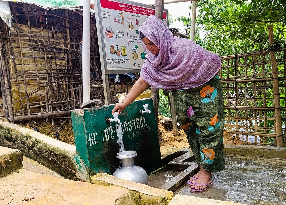 Eine Frau steht draußen an einer Wasserentnahmestelle und lässt Wasser in einen Blechkrug laufen
