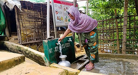 Eine Frau steht draußen an einer Wasserentnahmestelle und lässt Wasser in einen Blechkrug laufen