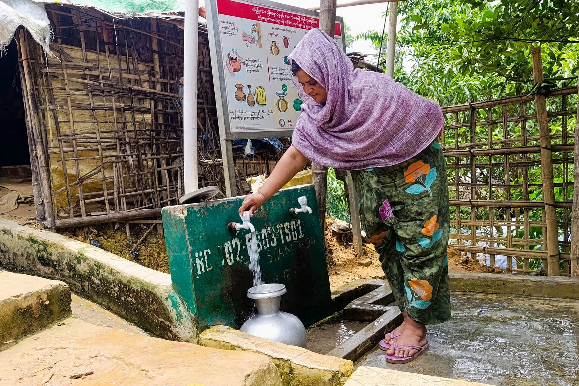 Eine Frau steht draußen an einer Wasserentnahmestelle und lässt Wasser in einen Blechkrug laufen