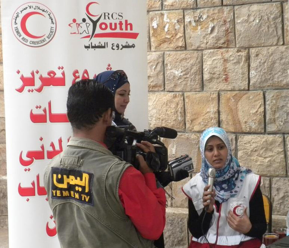 vor einem arabisch beschriebenen Plakat steht eine junge Muslima mit einem Mikrofon