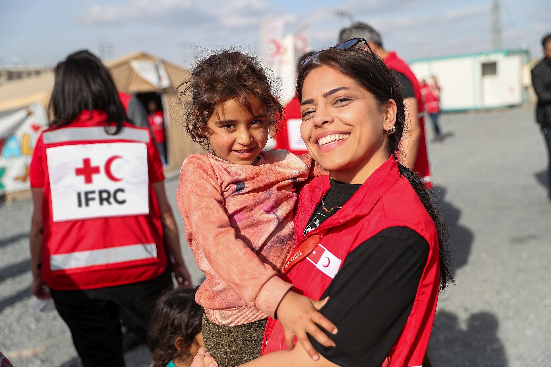 Eine Helferin des Türkischen Roten Halbmonds mit einen jungen Mädchen auf dem Arm. Beide lächeln in die Kamera.