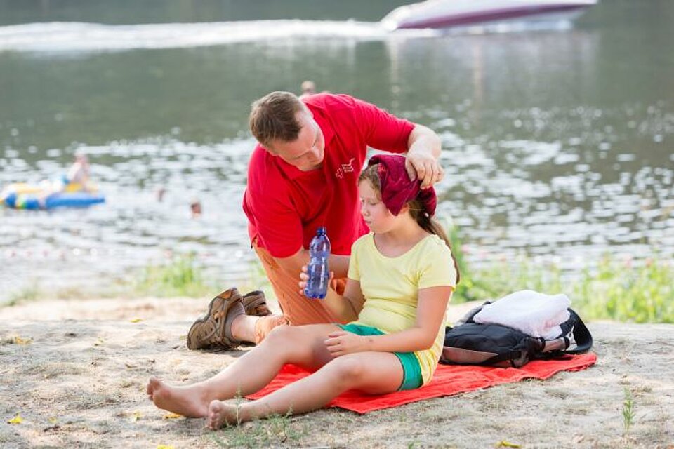 Ein Erst-Helfer versorgt ein Mädchen in der Hitze am Strand mit Wasser und kalten Umschlägen.