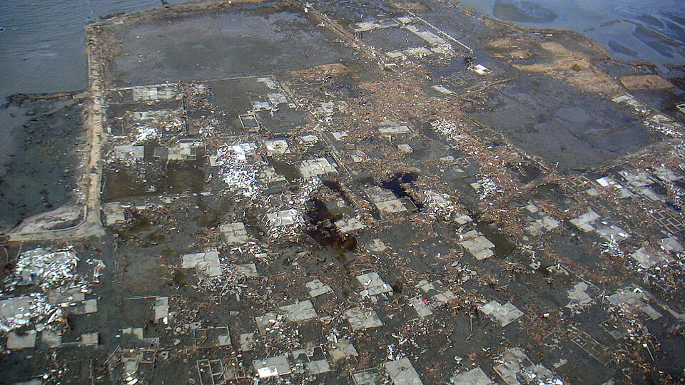Luftbild einer Küstenregion, die nach dem Tsunami 2004 vollständig zerstört wurde. Die Überreste von Gebäudefundamenten sind sichtbar, umgeben von Schlamm, Trümmern und Wasseransammlungen.
