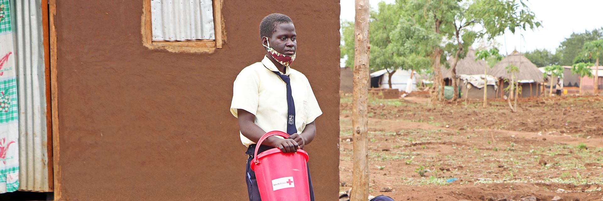 Eine ugandische Schülerin mit DRK-Eimer vor einem Haus
