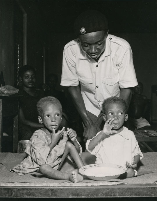 Foto: Ein nigerianischer Rotkreuzhelfer beugt sich über zwei essende Kleinkinder