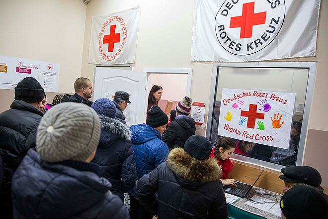Viele Menschen stehen vor einem Tisch. Eine Mitarbeiterin des Ukrainischen Roten Kreuzes arbeitet dort am Laptop. An der Wand hängen Banner des Ukrainischen Roten Kreuzes und des DRK.