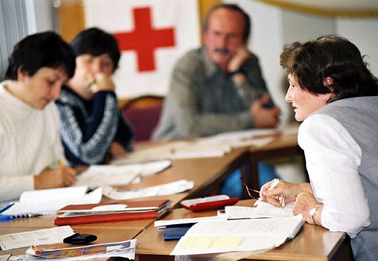 Foto: Ein Frau sitzt an einem Tisch mit einer Arbeitsgruppe, vor ihr ein Stapel Arbeitsblätter; unscharf im Hintergrund ein Mann und zwei weitere Frauen sowie ein Rotes Kreuz